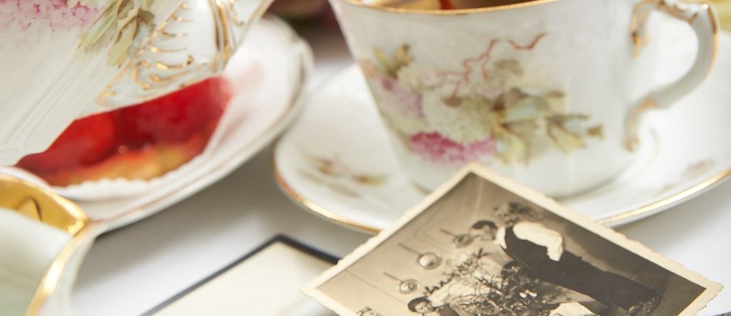 Ein Teeservice mit einem Teekessel, der Tee in eine Tasse mit Blumenmuster auf einer Untertasse gießt, umgeben von alten Fotos und einem Obstteller.