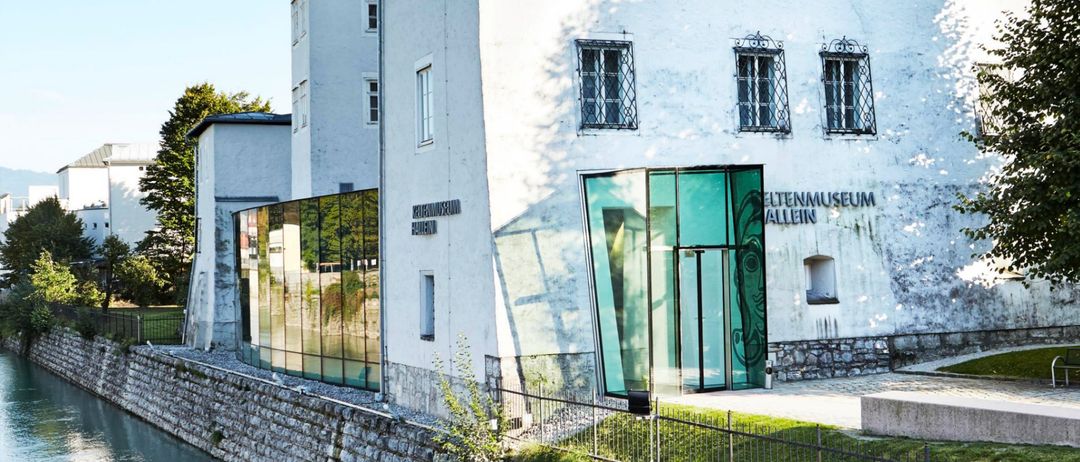 Eine moderne Glaserweiterung des weiß getünchten Museums Aller in Salzburg, Österreich, steht neben einem Kanal mit einer Steinmauer und einem Baum.