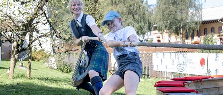 Zwei Personen, eine Frau und ein Kind, sind in einem freundschaftlichen Tauziehen auf einem Rasenfeld mit Bäumen und Gebäuden im Hintergrund.
