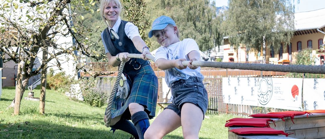 Zwei Personen, eine Frau und ein Kind, sind in einem freundschaftlichen Tauziehen auf einem Rasenfeld mit Bäumen und Gebäuden im Hintergrund.