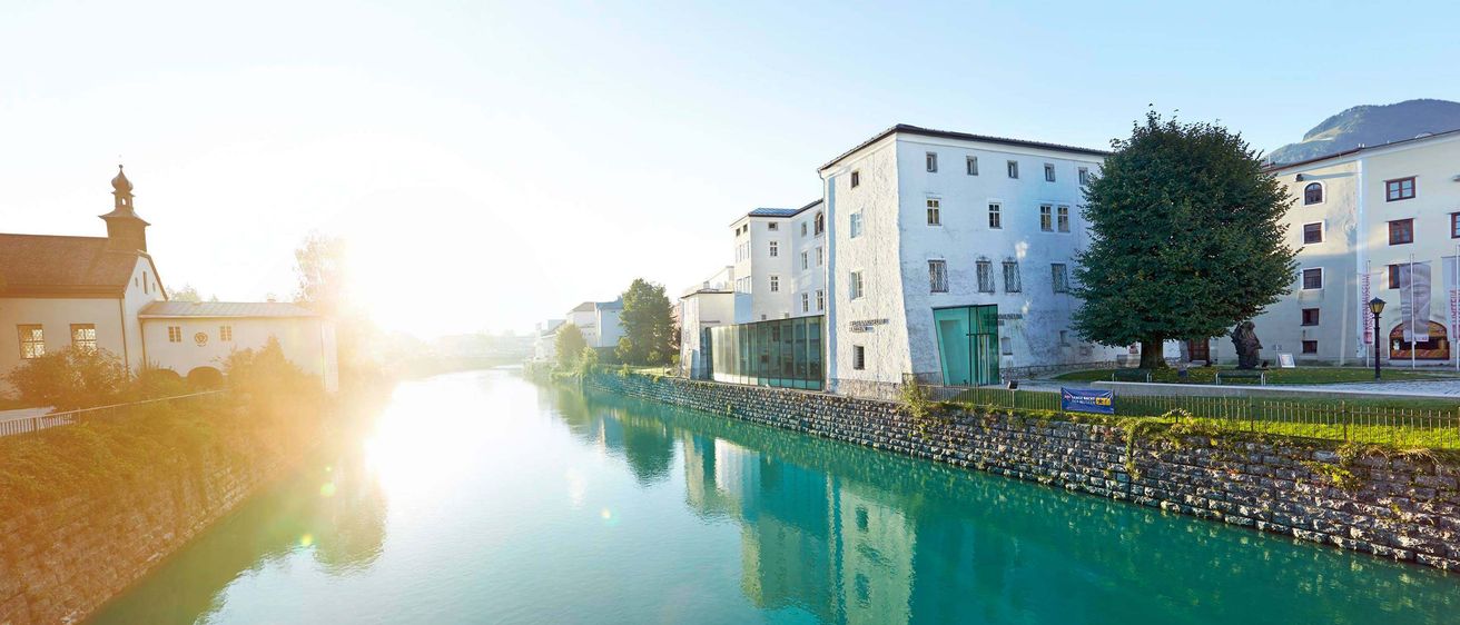 Eine ruhige Szene mit einem Fluss, der ein weißes Gebäude reflektiert, umgeben von Bäumen und Gebäuden, unter einem hellen Himmel.