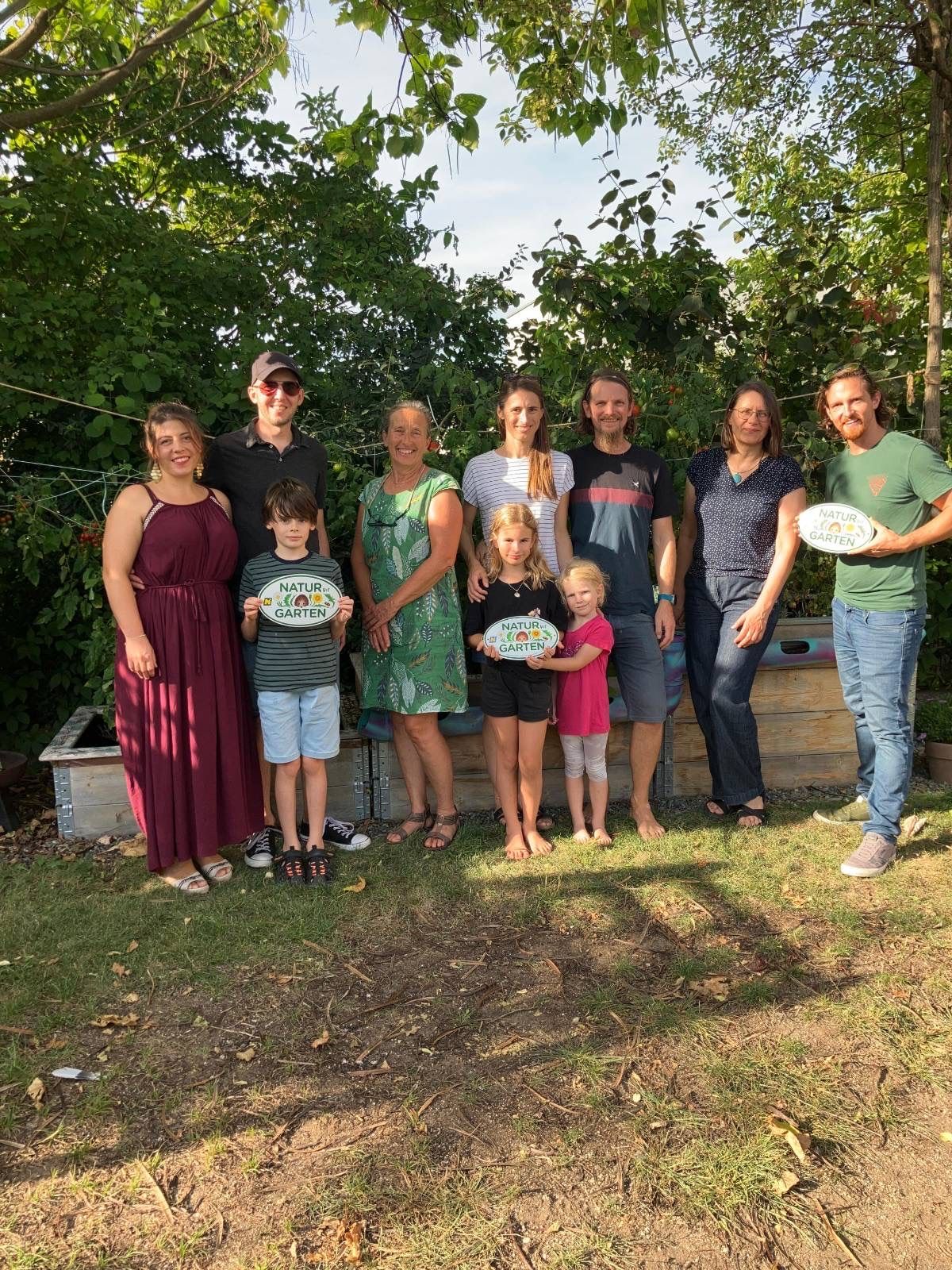 Eine Gruppe von Menschen steht in einem Garten, einige halten Schilder mit der Aufschrift 'Natur Garten'. Sie lächeln und posieren für ein Foto.
