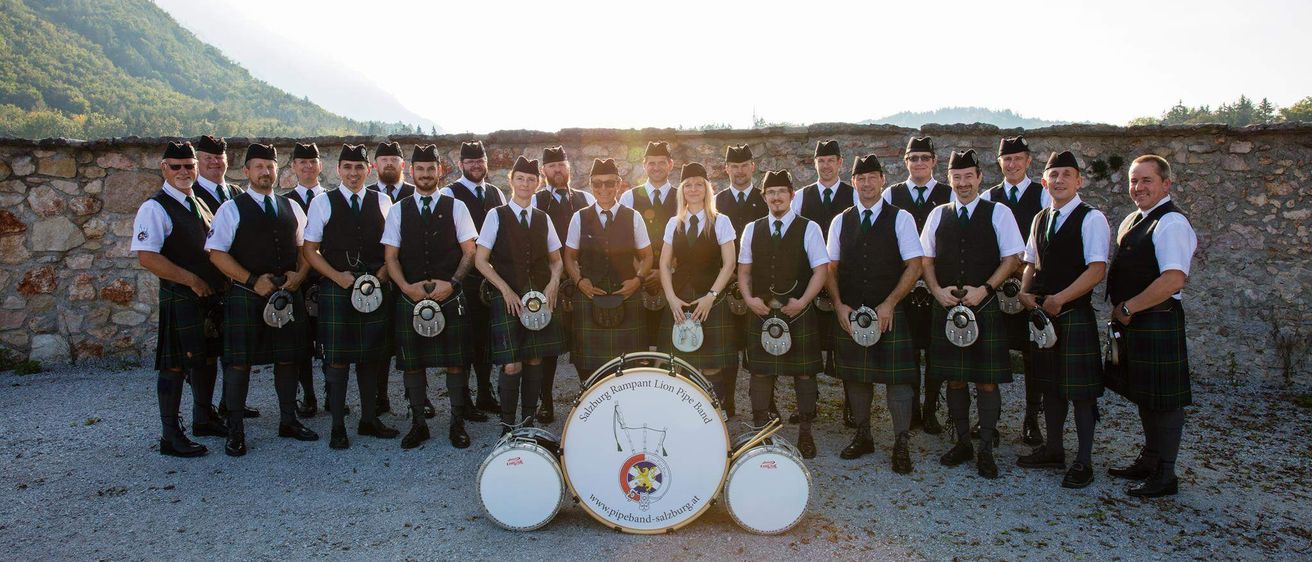 Eine Gruppe von Menschen in Kilts und Westen, die vor einer Steinmauer mit Trommeln und Dudelsack für ein Foto posieren.