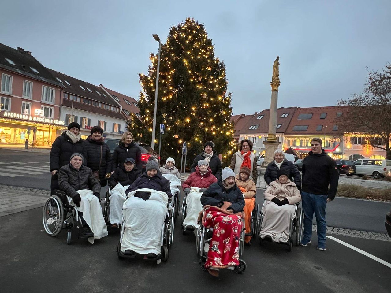 Eine Gruppe von Menschen in Rollstühlen, in Decken gewickelt, versammelt sich vor einem Weihnachtsbaum. Dahinter befinden sich mehrere Gebäude und eine Statue.