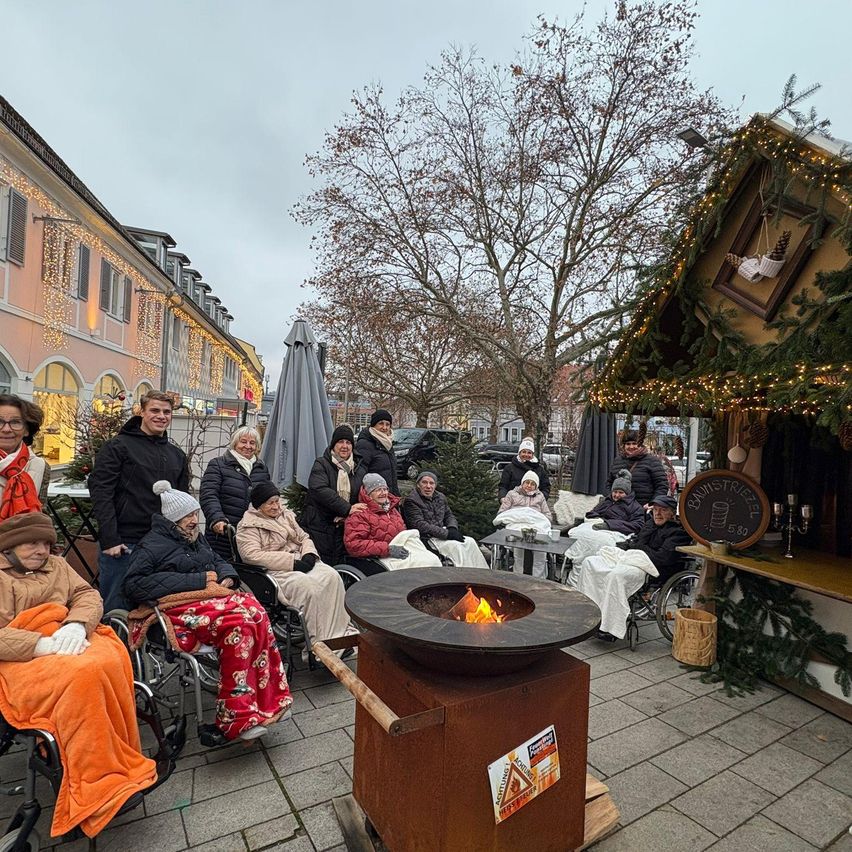 Eine Gruppe älterer Menschen in Rollstühlen sitzt um ein Feuer herum, mit einem für Weihnachten geschmückten Gebäude im Hintergrund.