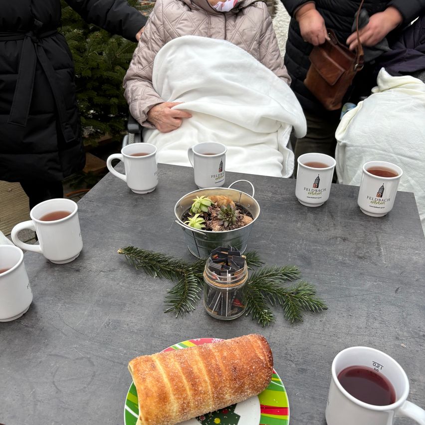 Ein Tisch mit Tassen Tee, einer Schüssel mit Essen und einem Teller mit einem Croissant auf einem bunten Teller. Eine ältere Person sitzt in einem Stuhl, eingewickelt in eine Decke.