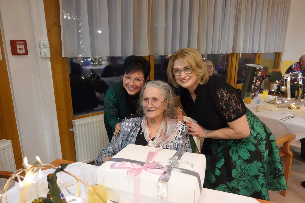 Drei Frauen lächeln für ein Bild. Die ältere Frau sitzt in der Mitte, mit einem Geschenkkarton vor ihr. Zwei Frauen stehen hinter ihr, von denen eine sie umarmt.