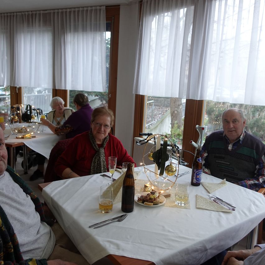 Mehrere ältere Menschen sitzen in einem Restaurant um einen Tisch herum, auf dem sich eine Weihnachtsbaumdekoration befindet. Der Tisch ist mit einer weißen Tischdecke bedeckt, und darauf stehen Gläser und ein Teller mit Essen. Das Restaurant hat Glasfenster mit weißen Vorhängen.
