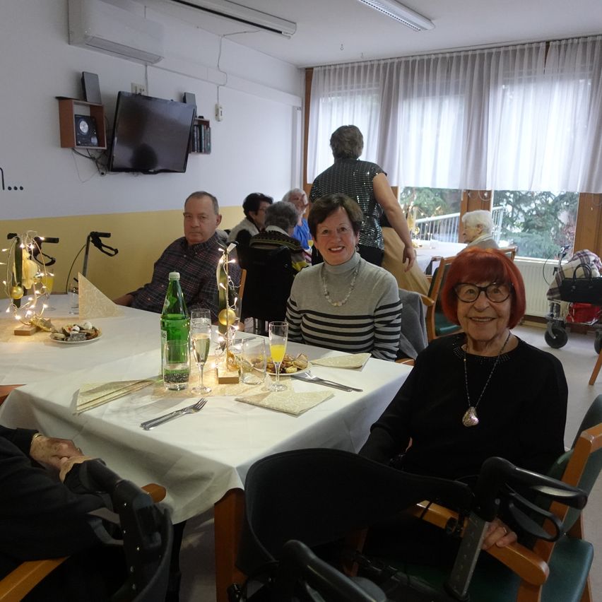 Mehrere Personen sitzen um einen Tisch in einem Raum. Der Tisch ist mit einem weißen Tuch bedeckt und hat verschiedene Gegenstände darauf. Eine Frau mit Brille sitzt in einem Stuhl mit Rollator.