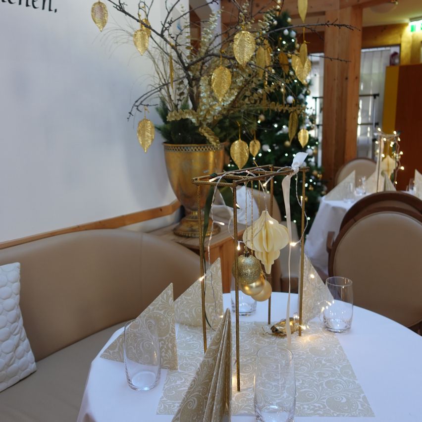 Ein Tisch für das Essen in einem Restaurant mit einem Weihnachtsbaum und Dekorationen im Hintergrund. Der Tisch hat eine weiße Tischdecke, Gläser und eine Serviette mit einem dekorativen Ornament.