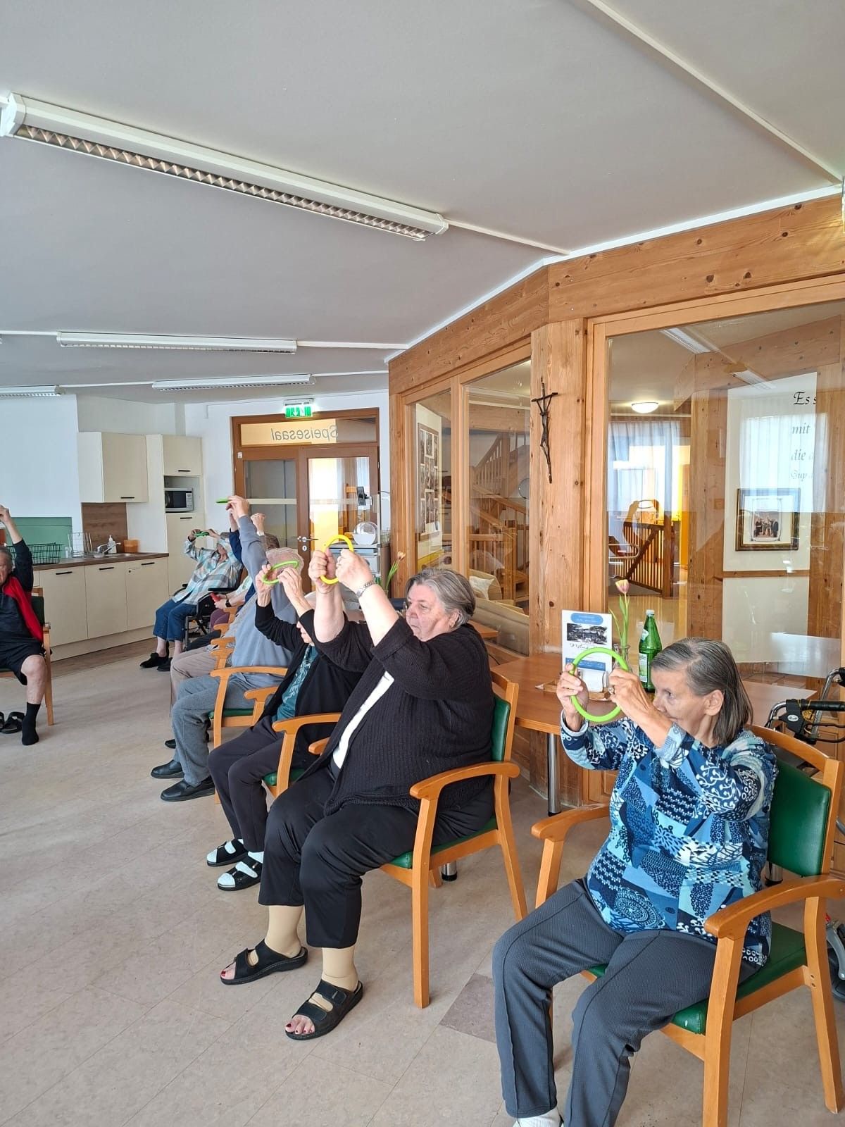 Eine Gruppe älterer Personen sitzt in Stühlen in einem Raum, einige tragen Brillen und halten grüne Ringe. Der Raum hat Holzwände und Glastüren, ein Treppenhaus und ein Küchenbereich sind sichtbar.