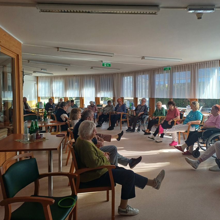 Eine Gruppe älterer Menschen sitzt in Stühlen um einen Raum herum, einige in Rollstühlen, mit Tischen und Flaschen darauf, und weiße Vorhänge bedecken die Fenster.