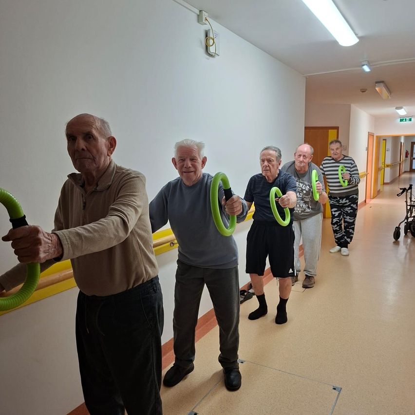 Fünf ältere Männer trainieren in einem Flur mit gelben Handläufen. Sie halten grüne Übungsbänder.