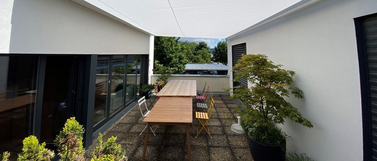 Eine Terrasse mit einem weißen Vordach, Holztischen und bunten Stühlen. Die Terrasse hat einen Kiesboden und eine weiße Wand mit Topfpflanzen. In der Ferne gibt es Bäume und ein Gebäude.