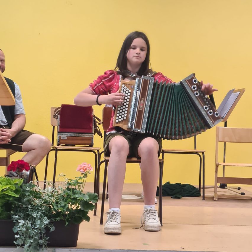 Ein Mädchen spielt auf der Bühne ein Akkordeon, mit einer gelben Wand im Hintergrund und zwei Männern, die auf Stühlen sitzen.