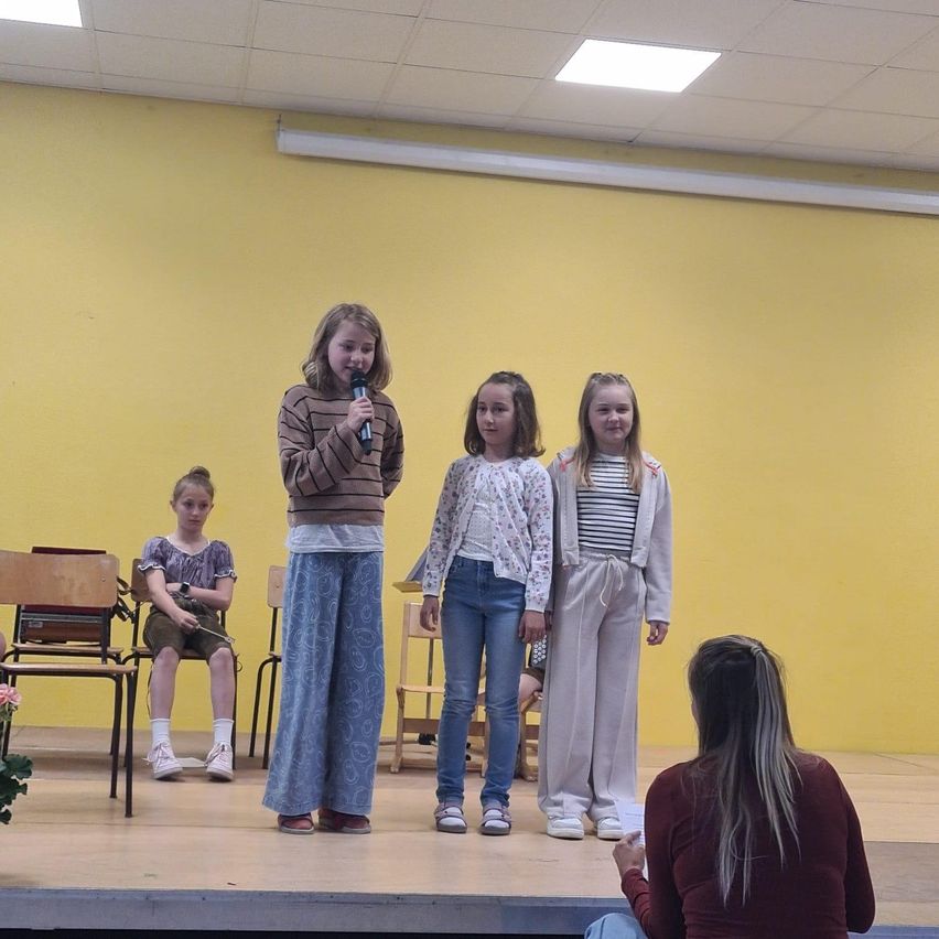 Vier Mädchen auf der Bühne, eine mit Mikrofon, drei stehend, eine sitzend, mit einer Frau, die von der Seite beobachtet.