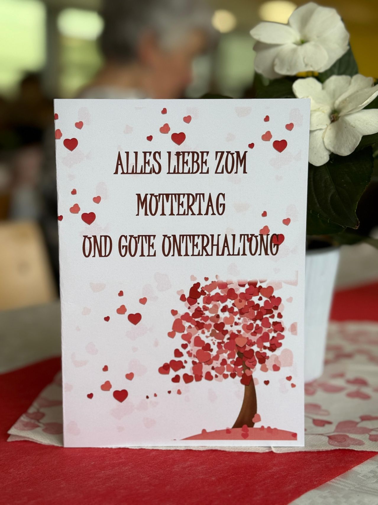 Eine Karte mit einem Herzbaum-Design, 'Alles Liebe zum Muttertag und gute Unterhaltung' in fetter roter Schrift. Eine weiße Blume in einem Topf ist im Hintergrund verschwommen.