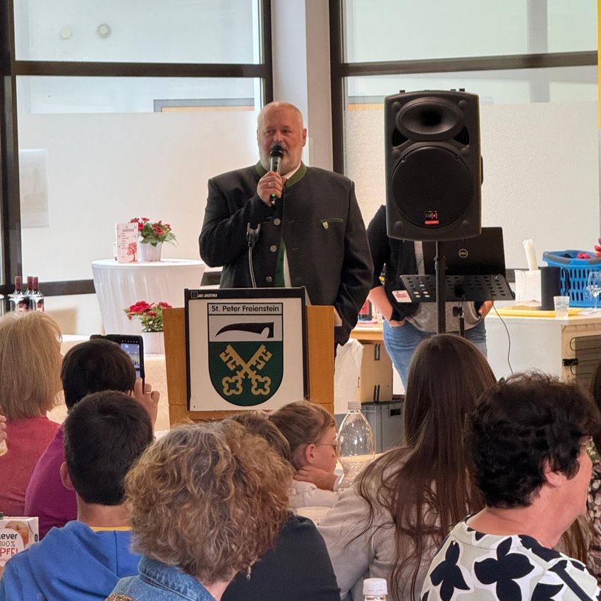 Ein Mann spricht in ein Mikrofon an einem Podium mit einem Schild davor, umgeben von einem Publikum, mit einem Lautsprecher und einem Laptop dahinter.