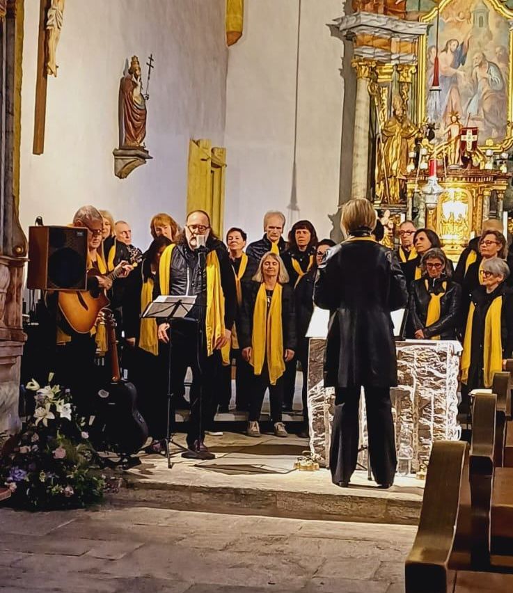 Ein Chor von Erwachsenen, die gelbe Schals tragen, tritt in einer Kirche mit Mikrofon, Lautsprecher und Gitarre auf.