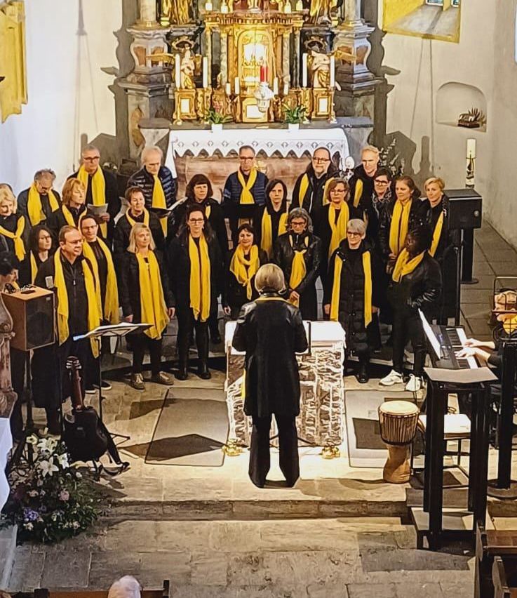 Ein Chor steht in einer Kirche und trägt gelbe Schals. Eine Person spielt Klavier, während ein Schlagzeuger ein Schlagzeug spielt.