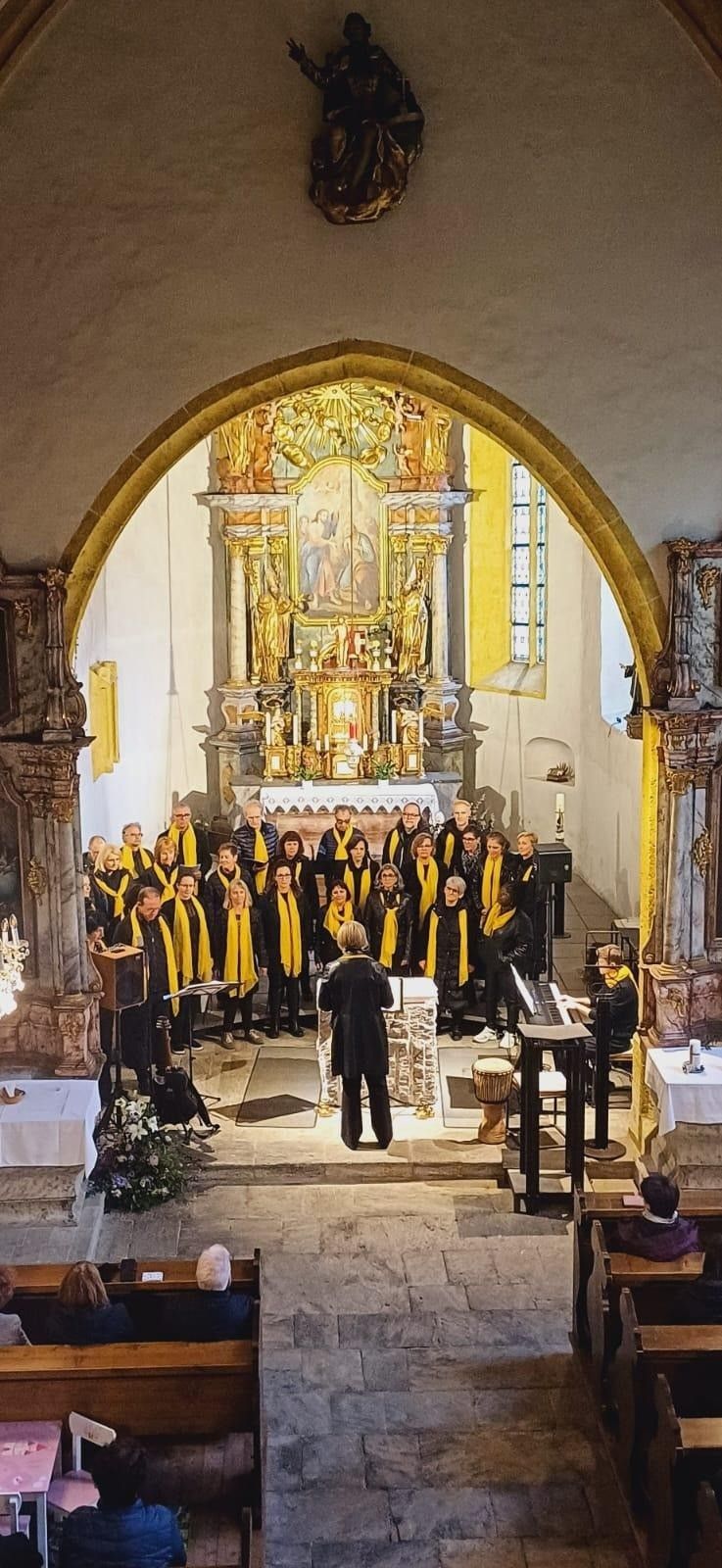 Ein Chor in gelben Schals singt in einer Kirche, mit einem Dirigenten und einem Pianisten. Ein Altar mit einem Gemälde und Kerzen im Hintergrund.