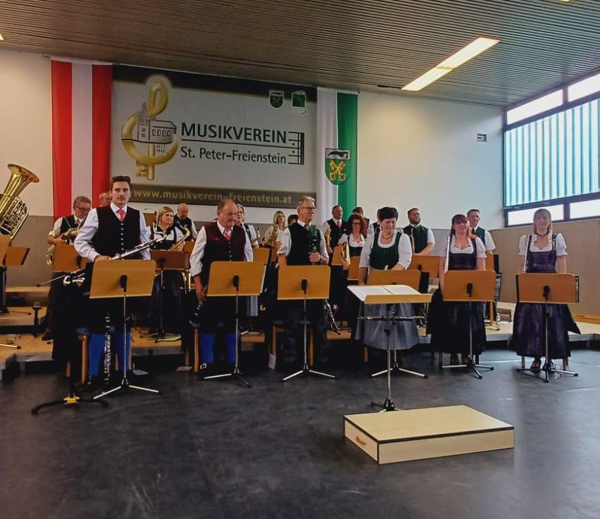 Eine Musikband tritt in einem Raum mit Bannern und Flaggen auf. Sie stehen vor Notenständern mit Instrumenten.