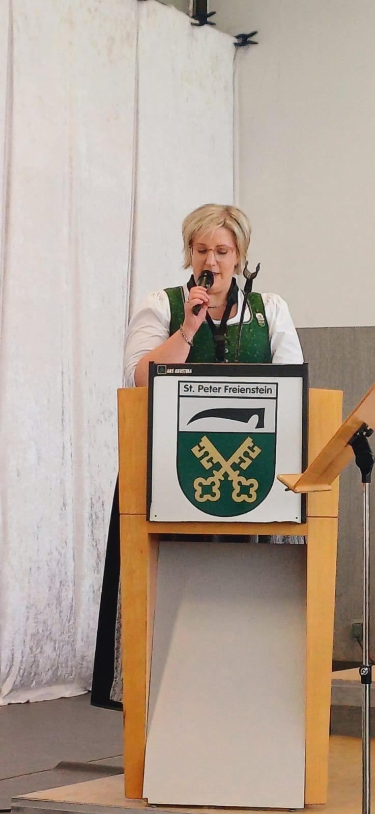 Eine Frau spricht in ein Mikrofon an einem Podium mit einem Schild, das St. Peter Freienstein steht. Sie trägt eine Brille und eine grüne Schürze. Hinter ihr befindet sich ein weißer Vorhang.