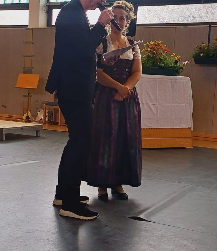 Ein Mann und eine Frau stehen auf einer Bühne; der Mann hält ein Mikrofon und Papier, und die Frau hält ein Stück Papier. Hinter ihnen steht ein Tisch mit weißer Tischdecke und Pflanzen.