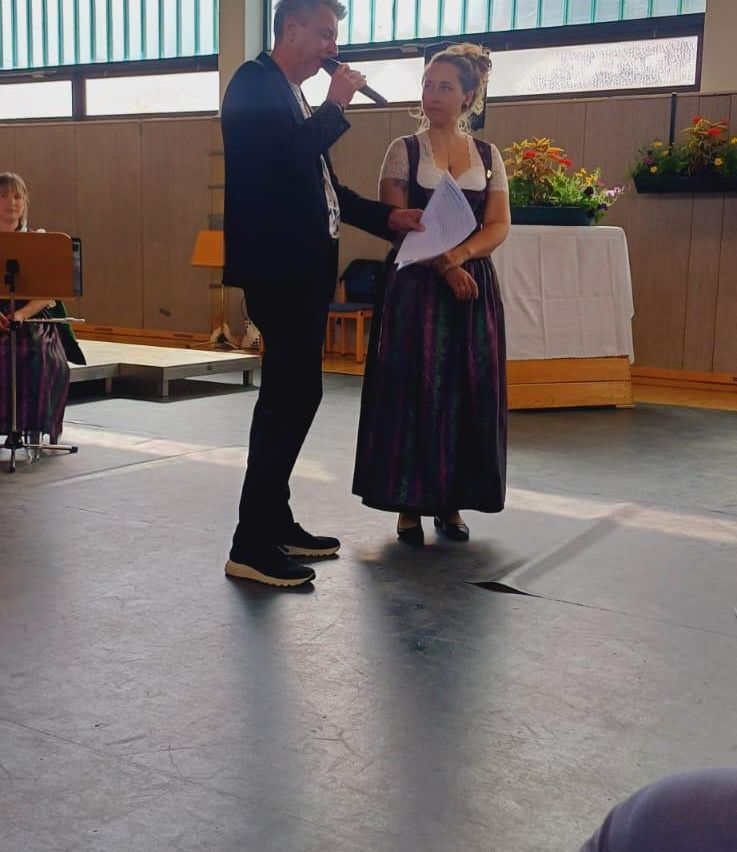 Ein Mann mit einem Mikrofon spricht zu einer Frau in traditioneller Kleidung, die auf einer Bühne steht, mit einem Tisch und Blumen hinter ihnen.