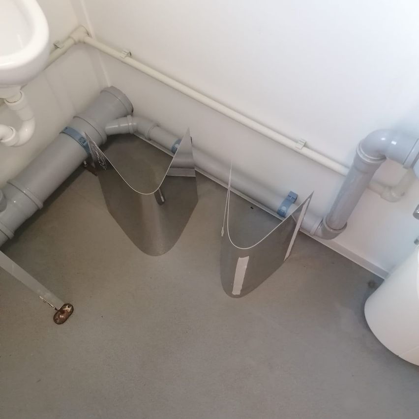 Ein Badezimmerboden mit weißen Fliesen und Metallrohren. Ein Waschbecken und eine weiße Toilette sind sichtbar. Es gibt zwei graue Schutzbarrieren.