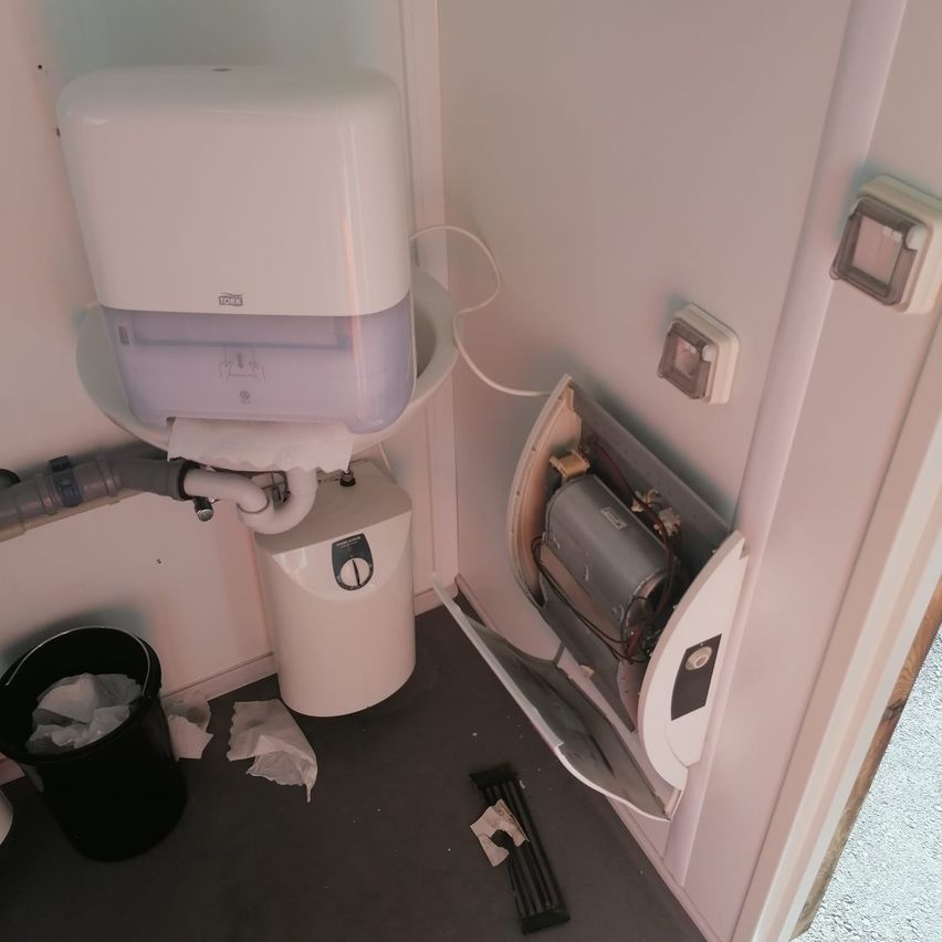 Ein kleines Badezimmer mit einem Toilettenpapierspender, einem Warmwasserbereiter und einer offenen Schalttafel an der Wand. Ein schwarzer Mülleimer steht auf dem Boden.