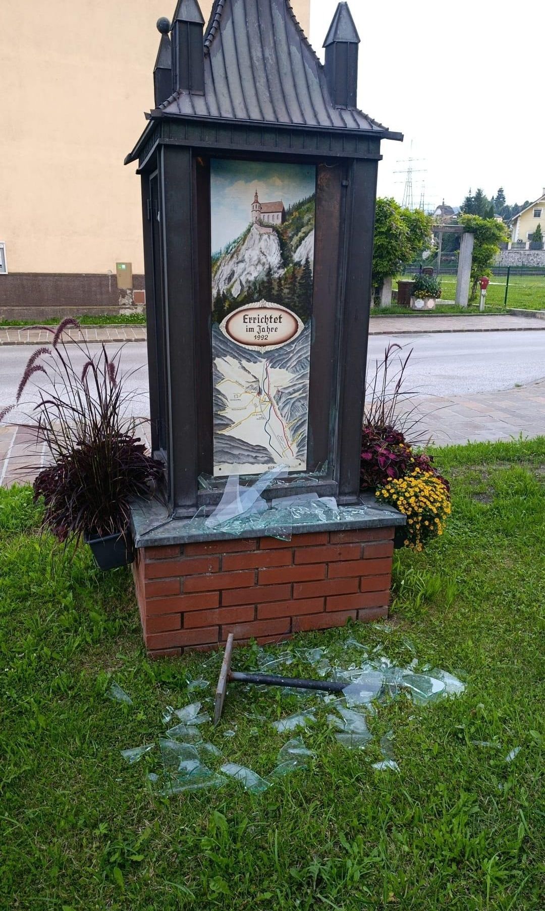 Eine Struktur aus Ziegelsteinen mit zerbrochenem Glas und einer Karte darauf steht auf einem Rasen. Es hat zwei Topfpflanzen auf beiden Seiten. Die Struktur hat den Text Errichtet im Jahre 1992 darauf.