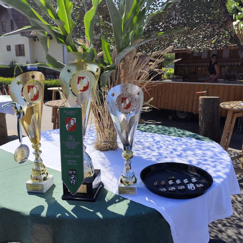 Drei Trophäen sind auf einem Tisch mit grüner Tischdecke ausgestellt. Sie sind von Medaillen und einer schwarzen Schale umgeben. Eine Pflanze steht auf dem Tisch. Hinter dem Tisch steht eine Frau vor einem Imbisswagen.