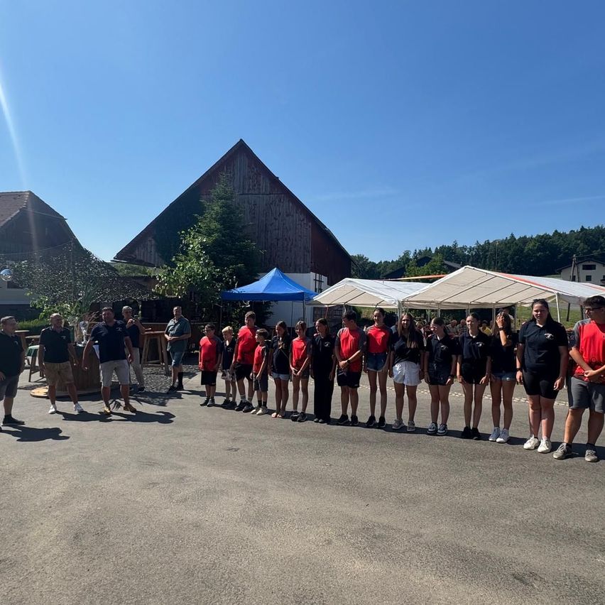 Eine Gruppe von Menschen steht in zwei Reihen in einer ländlichen Umgebung. Sie tragen passende rote und schwarze Uniformen. Hinter ihnen befinden sich Zelte und eine Scheune, mit Bäumen und Bergen in der Ferne.