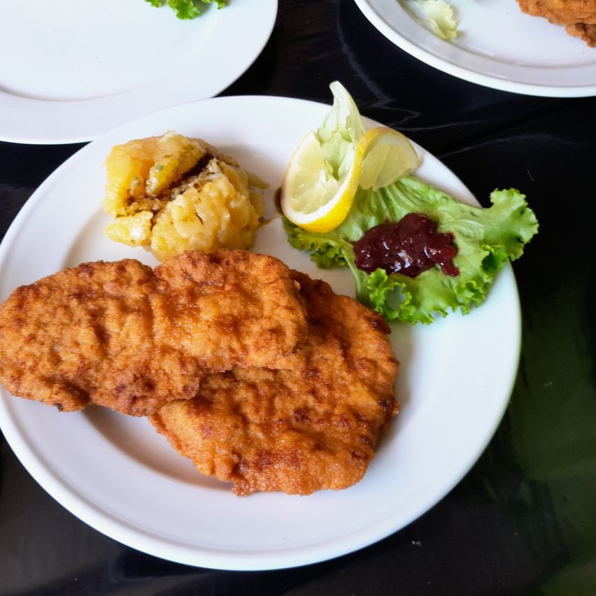 Ein Teller mit gebratenem paniertem Fleisch, Kartoffelpüree und einer Beilage Salat mit Zitrone und Marmelade. Ein weiterer Teller im Hintergrund.