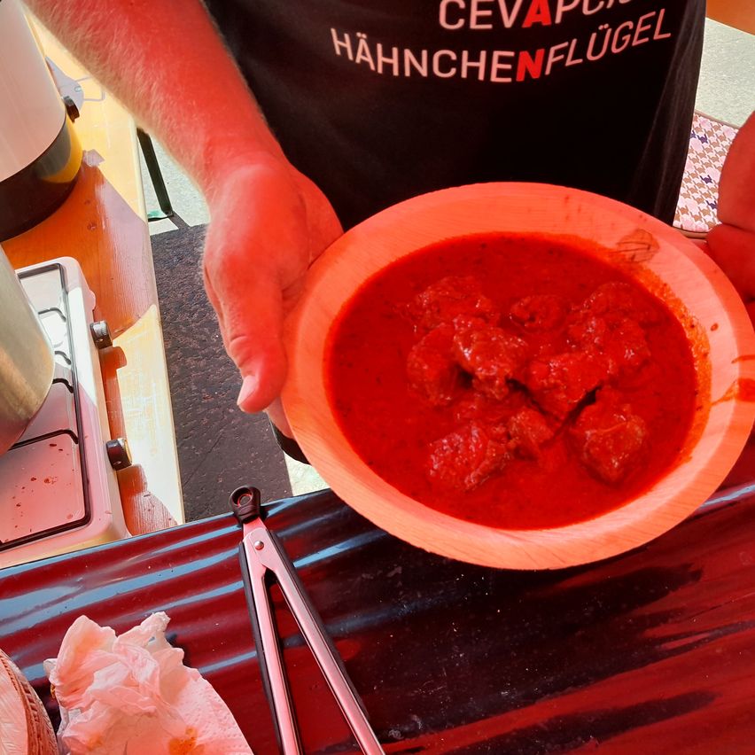 Eine Person, die ein Shirt mit 'CEVAN HAHNCHENFLUG' trägt, hält eine Schüssel mit rotem Fleischeintopf auf einem Holztisch. Küchenutensilien und ein Herd sind im Hintergrund zu sehen.
