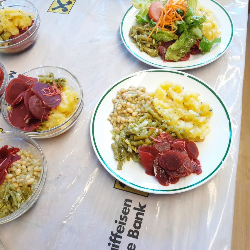 Ein Tisch zeigt fünf Schüsseln und zwei Teller mit Essen. Jede Schüssel enthält verschiedene Zutaten wie Gemüse und Obst. Ein Teller hat einen Salat mit Grünzeug und einer Tomate. Ein anderer Teller kombiniert Bohnen, Kartoffeln und Rote Bete.