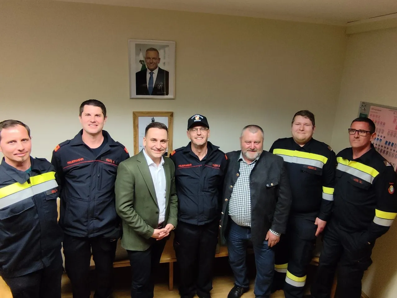 Sechs Feuerwehrleute in Uniform posieren mit einem Mann in einem grünen Anzug für ein Foto, stehen in einem Raum mit einem Bilderrahmen an der Wand.