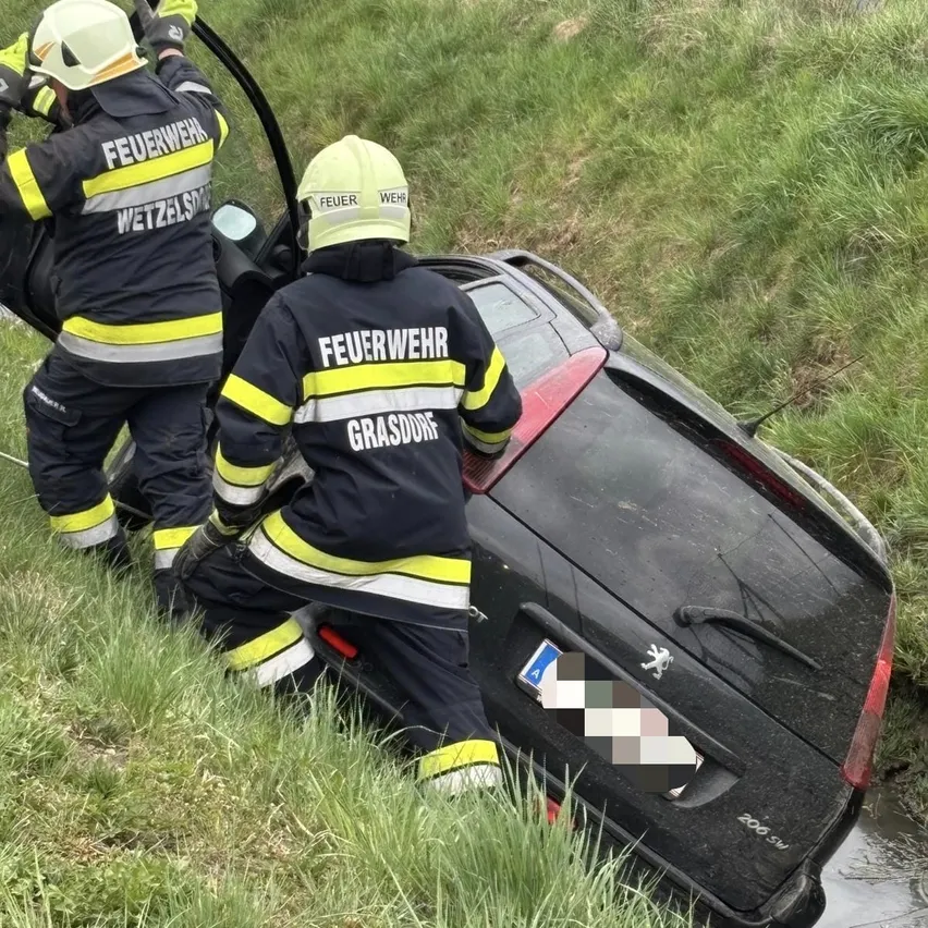 Zwei Feuerwehrleute versuchen, ein Auto aus dem Schlamm zu holen. Sie tragen Uniformen mit der Aufschrift 'Feuerwehr.' Das Auto steckt im Schlamm fest.