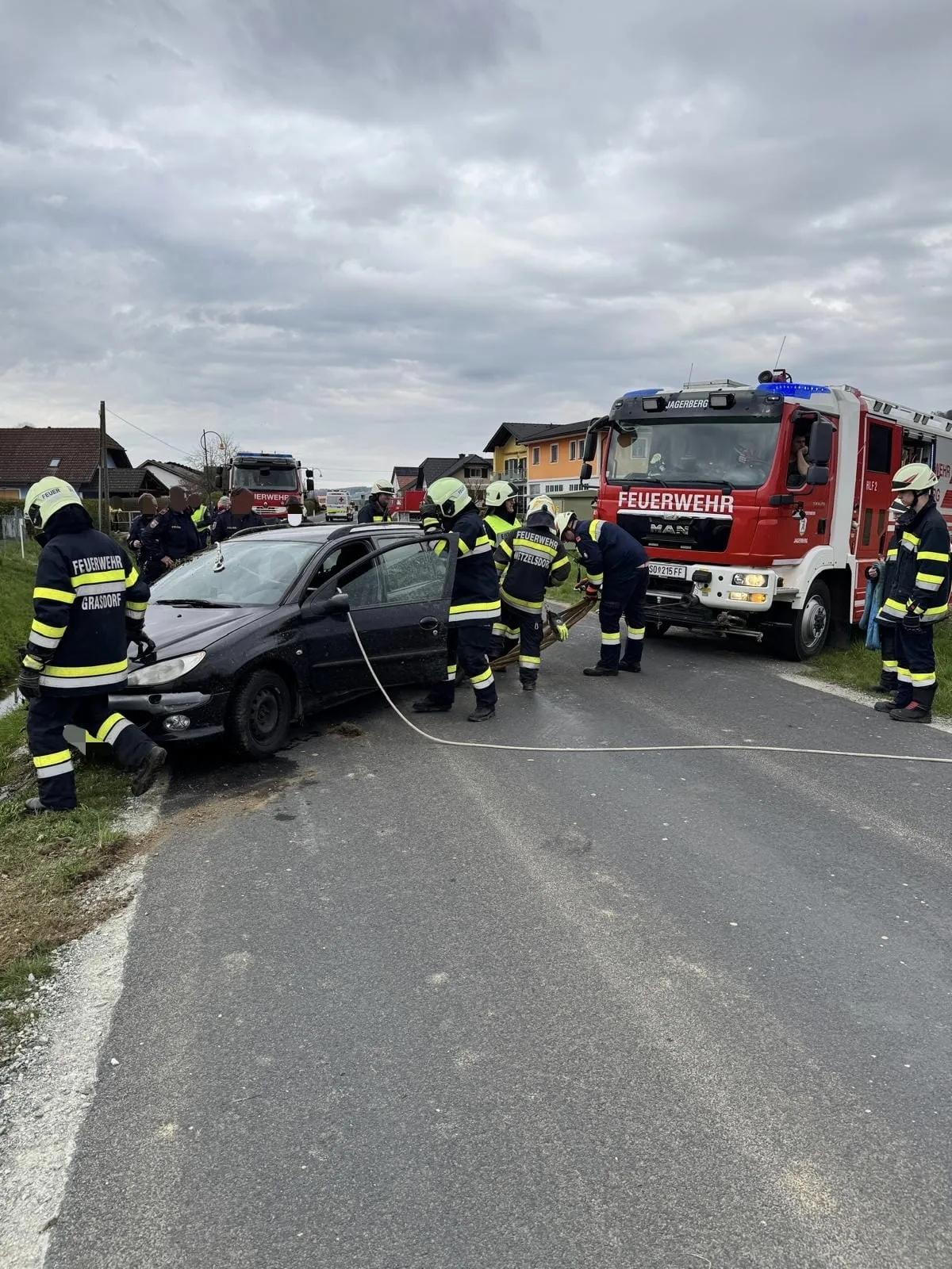 Ein schwarzer Wagen wird von Feuerwehrleuten am Straßenrand abgeschleppt. Ein roter Feuerwehrwagen ist in der Nähe geparkt.