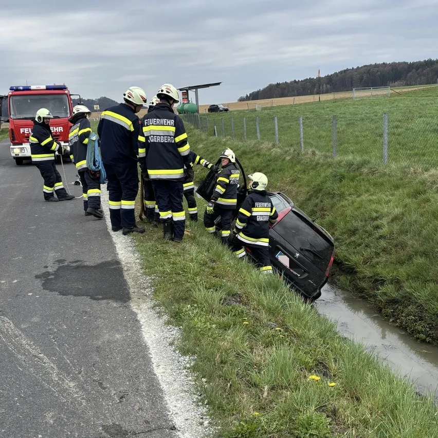 Feuerwehrleute stehen an einer Straße mit einem umgekippten Auto in einem Graben, ein Feuerwehrauto steht in der Nähe.