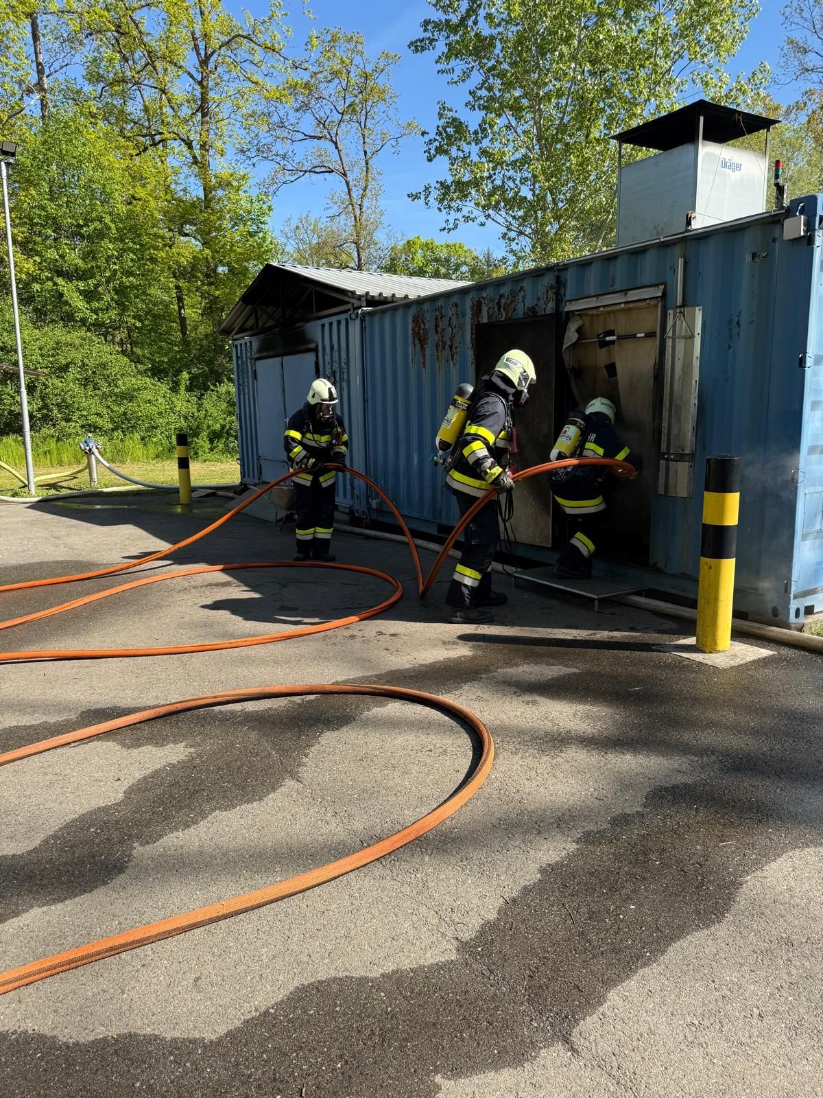 Feuerwehrleute üben mit einer Feuerwehrschlauch vor einem blauen Container, aus dem Rauch kommt. Im Hintergrund sind Bäume und Grünflächen zu sehen.