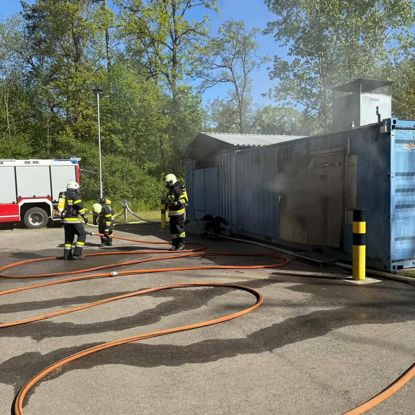 Feuerwehrleute arbeiten an einem blauen Container mit Rauch. Ein Feuerwehrauto ist in der Nähe geparkt.