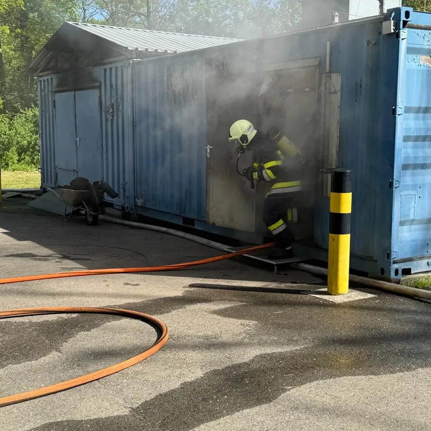 Ein Feuerwehrmann löscht einen Brand in einem blauen Container. Rauch kommt aus dem Container und eine Feuerwehrschläuche ist angeschlossen.