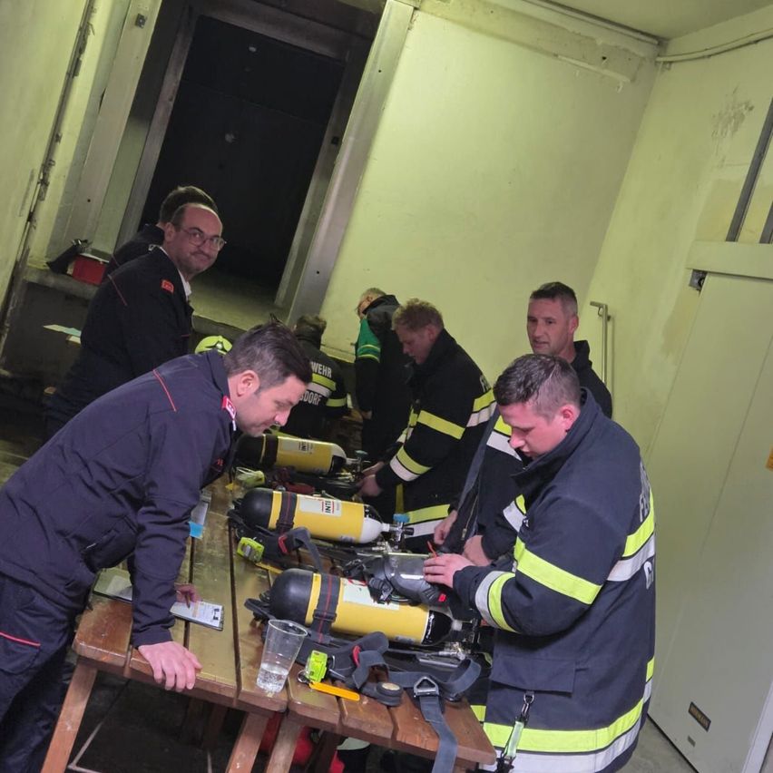 Fünf Feuerwehrleute stehen in einem Raum mit gelben Wänden um einen Tisch herum. Sie untersuchen und arbeiten an Ausrüstung, einschließlich Sauerstoffflaschen.