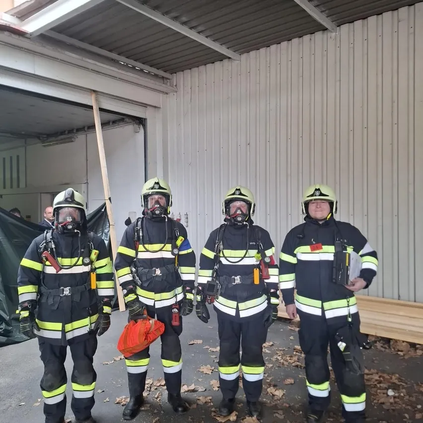 Vier Feuerwehrleute in voller Ausrüstung stehen in einer Garage, einer hält einen roten Beutel. Das Gebäude hinter ihnen hat weiße Wände und eine teilweise offene Garagentür.