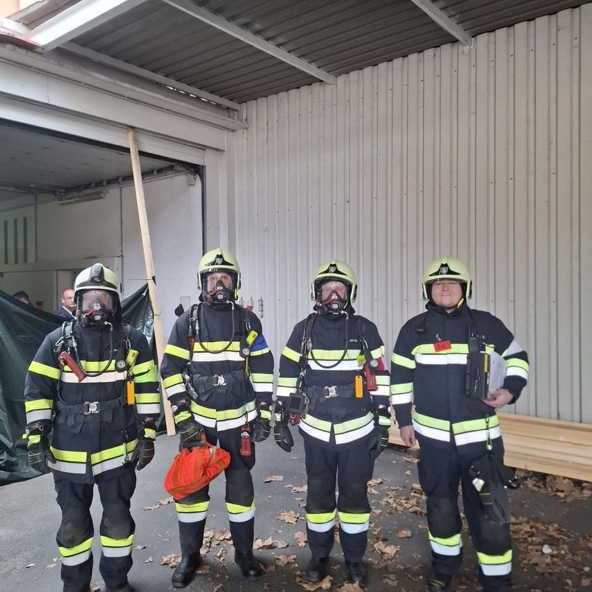 Vier Feuerwehrleute in voller Ausrüstung stehen in einer Garage, einer hält einen roten Beutel. Das Gebäude hinter ihnen hat weiße Wände und eine teilweise offene Garagentür.
