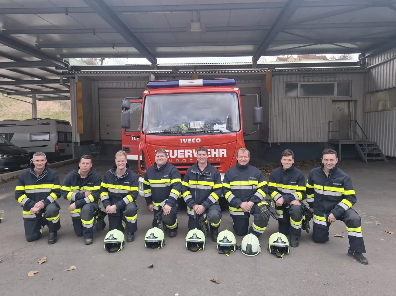 Eine Gruppe von Feuerwehrleuten posiert für ein Foto vor einem roten IVECO-Löschfahrzeug. Vor ihnen liegen Helme.