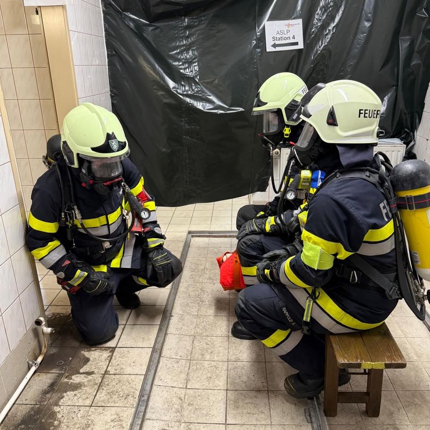 Drei Feuerwehrleute bereiten sich in einem Übungsbereich auf eine Rettungsaktion vor. Einer kniet auf dem Boden, ein anderer sitzt auf einer Bank und der dritte steht. Sie sind mit Helmen und Sauerstoffflaschen ausgestattet.