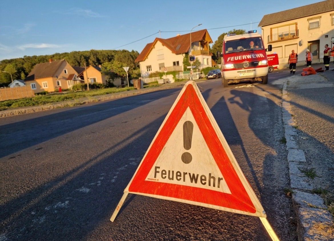 Ein rotes und weißes Warnschild für die Feuerwehr ist auf der Straße platziert, mit einem Feuerwehrauto im Hintergrund.